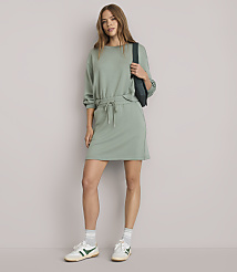 A woman in a light green sweatshirt dress and sneakers stands against a plain white background, with a dark green fanny pack slung across her chest.