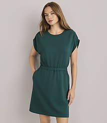 A woman in a green dress and sunglasses leans against a plain gray wall, with a light blue sweater tied around her waist. carousel Product Image 1
