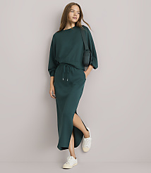 A woman in a teal sweatshirt and matching skirt with a side slit stands against a plain white background, wearing white sneakers and a small crossbody bag. carousel Product Image 1