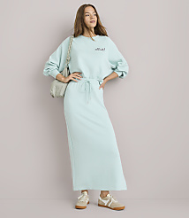A young woman in a light blue sweatshirt and matching maxi skirt stands with her hands on her hips against a plain white background, a light gray purse slung over her shoulder. carousel Product Image 1