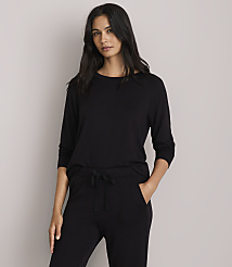 A woman in a black long-sleeved shirt and black pants stands against a plain gray background.