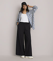 A woman in a white tank top, black wide-leg pants, and an open plaid shirt stands against a plain gray background. carousel Product Image 1