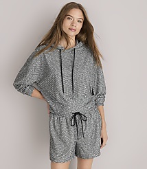 A woman in a grey patterned hoodie and shorts sits against a plain grey background.
