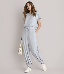 A woman in a light blue loungewear set and white sneakers stands against a plain white background, holding a cream-colored tote bag.