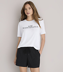 A woman in a white "LOW MAINTENANCE" t-shirt and black baseball cap poses against a plain white background. carousel Product Image 1