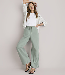 A woman in a white t-shirt, patterned shirt, and loose green pants stands against a plain white background. carousel Product Image 1