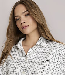 A young woman with long brown hair stands in the foreground wearing a checkered shirt and striped pants, against a plain white background. carousel Product Image 2