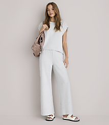 A young woman in a grey loungewear set and white sandals stands against a plain white background, with a pink backpack slung over her shoulder.