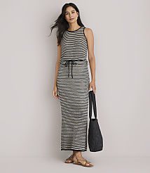A woman in a striped maxi dress and sandals stands against a plain white background, holding a black quilted tote bag.