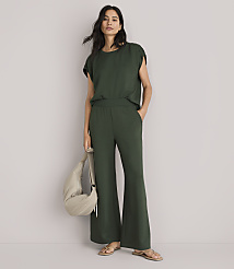 A woman in a green top and wide-leg pants stands against a plain white background, holding a beige bag.