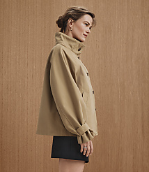 A woman in a tan jacket and black skirt stands against a warm, wood-paneled background. carousel Product Image 5