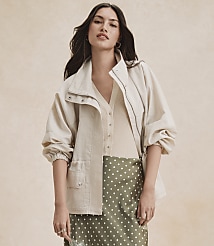 A woman in a cream jacket and green polka dot skirt stands against a textured beige background. carousel Product Image 4