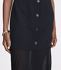 A close-up shot focuses on the buttoned front of a black dress with a sheer pleated skirt peeking out from the bottom, while a person's arm with a bracelet is visible on the left. carousel Product Image 4