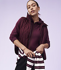 A woman in a quilted vest and hoodie stands against a plain purple background, holding a striped sweater. carousel Product Image 4