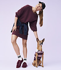 A young woman in a maroon dress and baseball cap poses with a small dog wearing a striped shirt against a light purple background. carousel Product Image 2