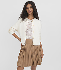 A woman in a white cardigan and tan pleated skirt stands against a plain white background. carousel Product Image 1