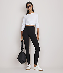 A woman in a white long-sleeved crop top and black leggings stands against a plain white background, holding a black bag. carousel Product Image 1