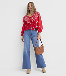 A woman in a red ruffled blouse and wide-leg jeans stands against a plain white background, holding a brown studded purse. carousel Product Image 2