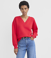A woman in a red v-neck sweatshirt and blue jeans stands against a plain white background. carousel Product Image 1