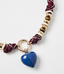 A blue heart charm hangs from a braided maroon and white cord necklace adorned with wooden and cream beads, set against a plain white background. carousel Product Image 1