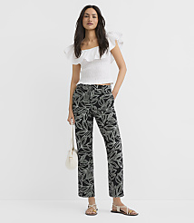 A woman in a white ruffled top and black leaf-patterned pants stands against a plain white background, holding a small white purse. carousel Product Image 1