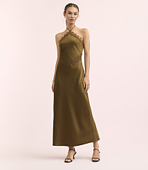 A woman in a long, olive-green halter dress with lace detailing stands against a soft pink background. carousel Product Image 1