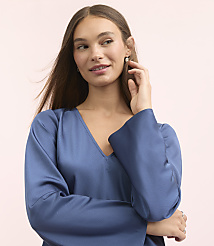 A young woman with long brown hair and a blue satin top poses with her hand near her ear against a soft pink background. carousel Product Image 4