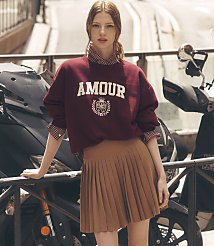 A young woman in a maroon sweatshirt and brown pleated skirt stands in front of a motorcycle with a blurred urban background. carousel Product Image 1