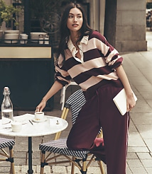 A woman in a striped shirt and maroon pants leans on a cafe table with a drink and book, while a blurred street scene with buildings and a sidewalk forms the background. carousel Product Image 1