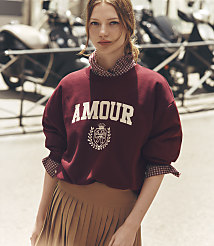 A young woman in a maroon sweatshirt with "AMOUR" written on it and a pleated skirt stands in front of a blurred background of parked motorcycles. carousel Product Image 1