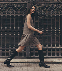 A woman in a plaid dress and black boots walks in front of an ornate metal gate. carousel Product Image 1