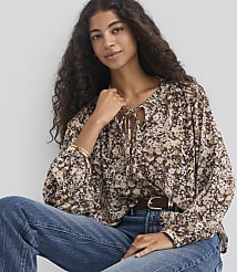 A woman with long, dark, curly hair wears a floral print blouse and blue jeans, seated against a plain, light-colored background. carousel Product Image 1