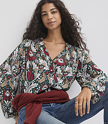 A woman with dark curly hair wears a dark green floral blouse and blue jeans, with a maroon sweater tied around her waist, against a plain white background. carousel Product Image 1