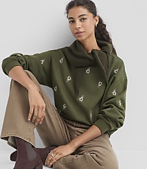A woman in a green embroidered sweatshirt and brown pants sits against a plain gray background. carousel Product Image 1