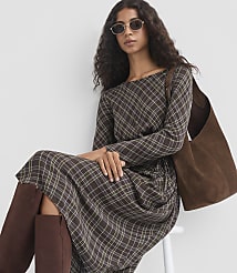 A woman in a plaid dress and brown boots sits in front of a white background, holding a brown suede bag. carousel Product Image 2