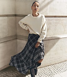 A woman in a cream sweater and plaid skirt stands against a textured stone wall. carousel Product Image 1
