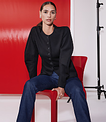 A woman in a black jacket and jeans sits on a red chair against a vibrant red backdrop with studio lighting equipment visible. carousel Product Image 4