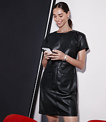 A woman in a black leather dress looks at her phone in the foreground, with a black and white background behind her. carousel Product Image 4