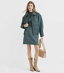 A woman in a teal jacket and skirt stands against a plain white background, holding a tan crossbody bag and wearing tan boots. carousel Product Image 1