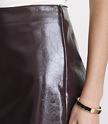 A close-up shot focuses on a shiny, burgundy faux leather skirt with a zipper detail, while a person's arm with a gold and black bracelet is visible on the right. carousel Product Image 4