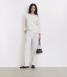 A woman in a cream sweater and matching pants stands against a plain white background, holding a small black purse. carousel Product Image 1