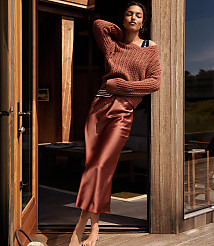 A woman in a rust-colored sweater and satin skirt leans against a wooden doorframe, with a blurred interior and outdoor scene in the background. carousel Product Image 4