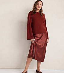 A woman in a rust-colored sweater and satin skirt stands against a plain, light-colored wall. carousel Product Image 1