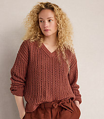 A young woman with curly blonde hair poses in a rust-colored, open-knit V-neck sweater and matching pants against a plain, light-colored wall. carousel Product Image 3