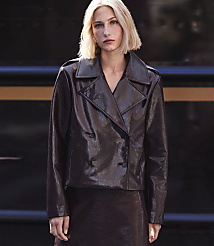 A blonde woman in a shiny brown jacket and skirt stands against a dark, textured background. carousel Product Image 6