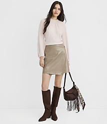 A woman in a light pink mock-neck top and a taupe faux-leather wrap skirt stands against a plain white background, accessorized with brown knee-high boots and a fringed crossbody bag. carousel Product Image 1