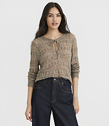 A woman in a light brown, open-knit sweater with a tie at the neck and dark wash jeans stands against a plain white background. carousel Product Image 2