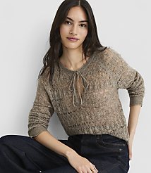 A woman in a sheer, taupe knit sweater with a tie-neck detail and dark wash jeans poses against a plain, light gray background. carousel Product Image 1