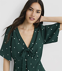 A woman in a green polka-dot dress with her hand on her head stands against a plain white background. carousel Product Image 1