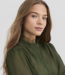A young woman with long, brown hair and blue eyes looks forward, wearing a green, high-necked blouse with lace trim, against a plain gray background. carousel Product Image 1